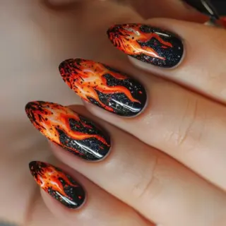 The nails are adorned with a black base and vivid flames that start from the tips. The flames transition from yellow at the tips to orange and red as they move towards the middle of the nails. The design includes small white sparkles, adding a touch of glamour to the fiery theme.
