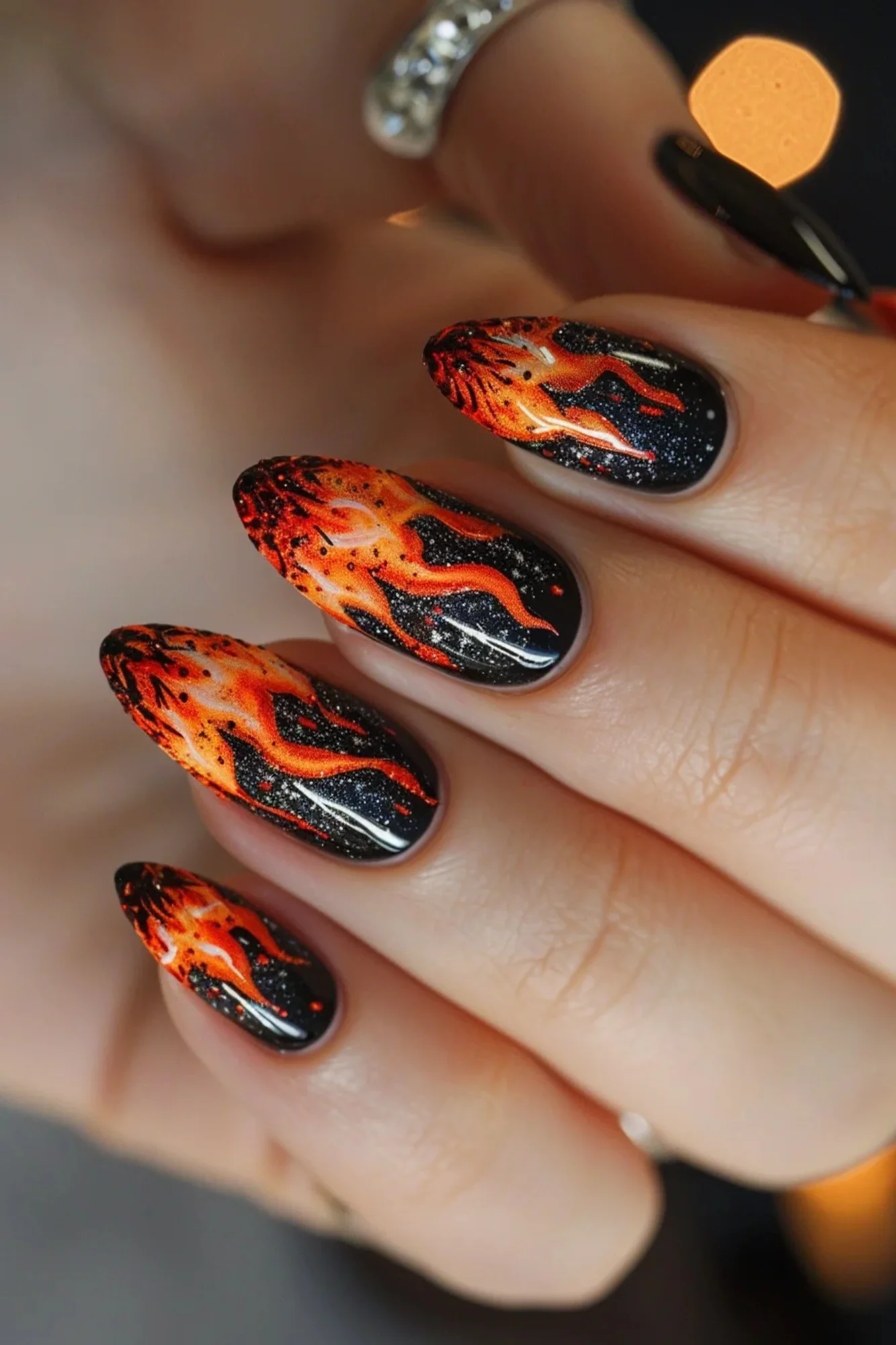 The nails are adorned with a black base and vivid flames that start from the tips. The flames transition from yellow at the tips to orange and red as they move towards the middle of the nails. The design includes small white sparkles, adding a touch of glamour to the fiery theme.