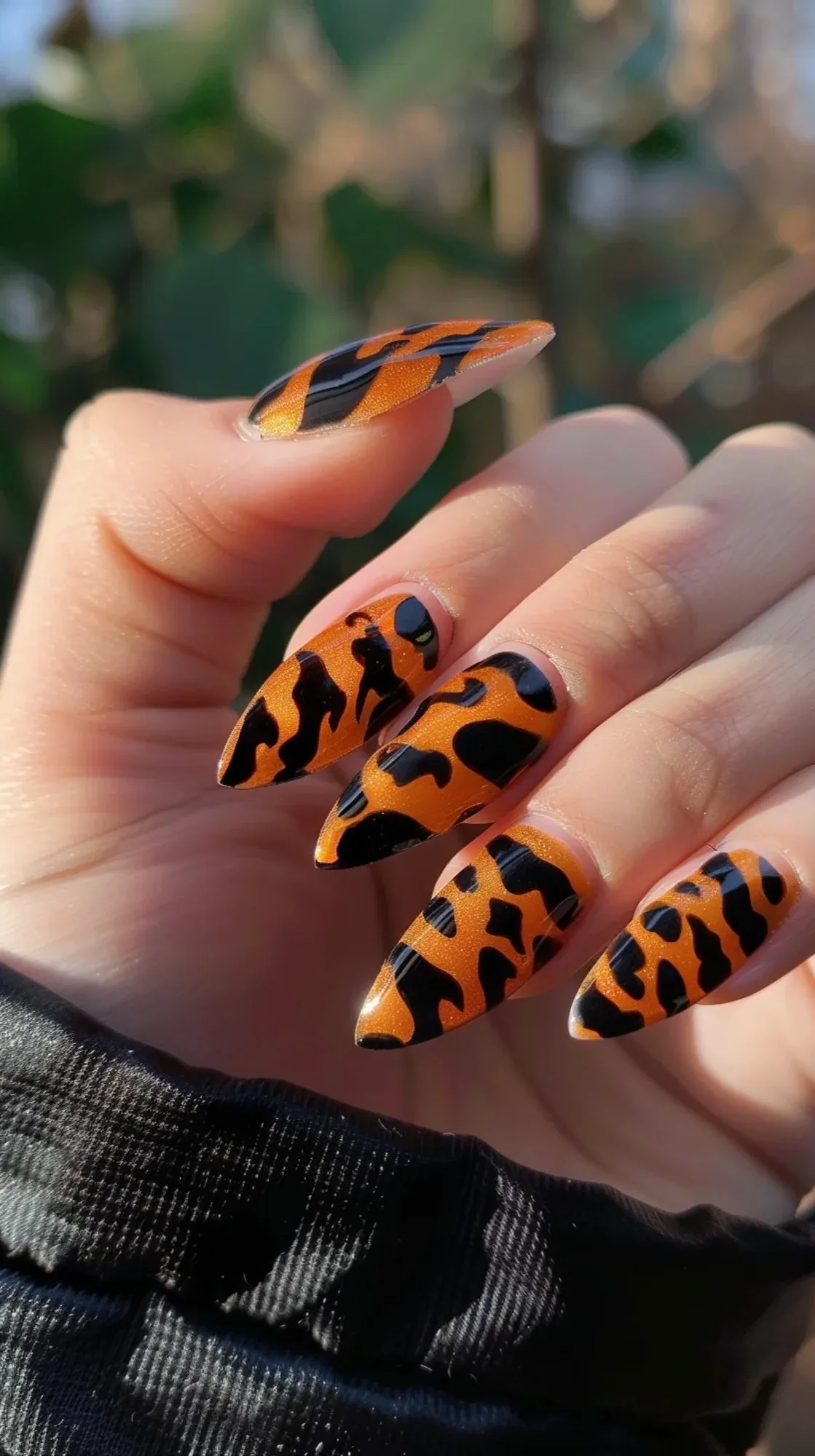 Long, almond-shaped nails feature a bold design with a shimmering orange base. The nails are adorned with a black tiger stripe pattern, creating a fierce and eye-catching look. 